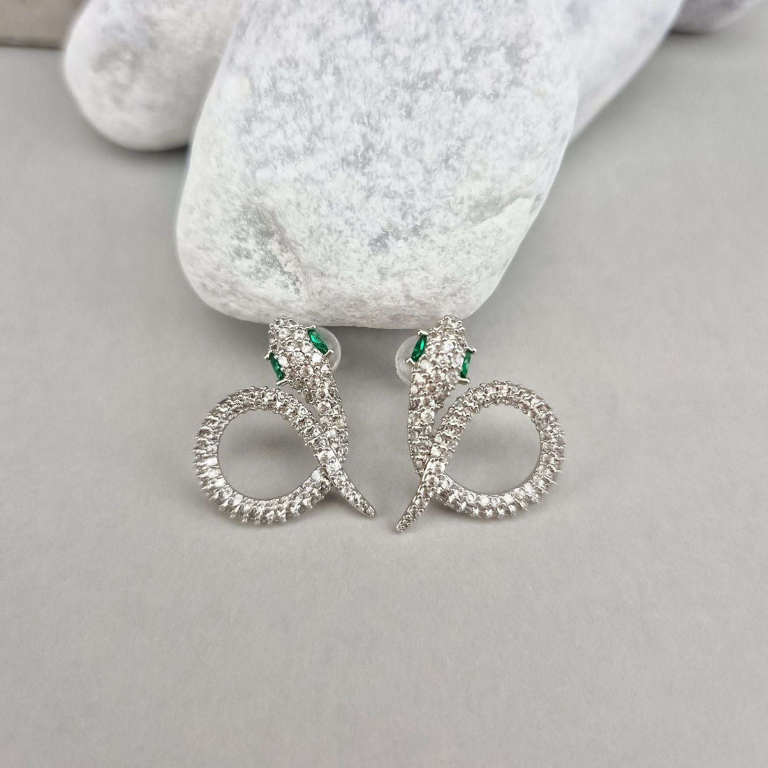Silver-tone snake earrings with clear stones and green eyes on a pale stone