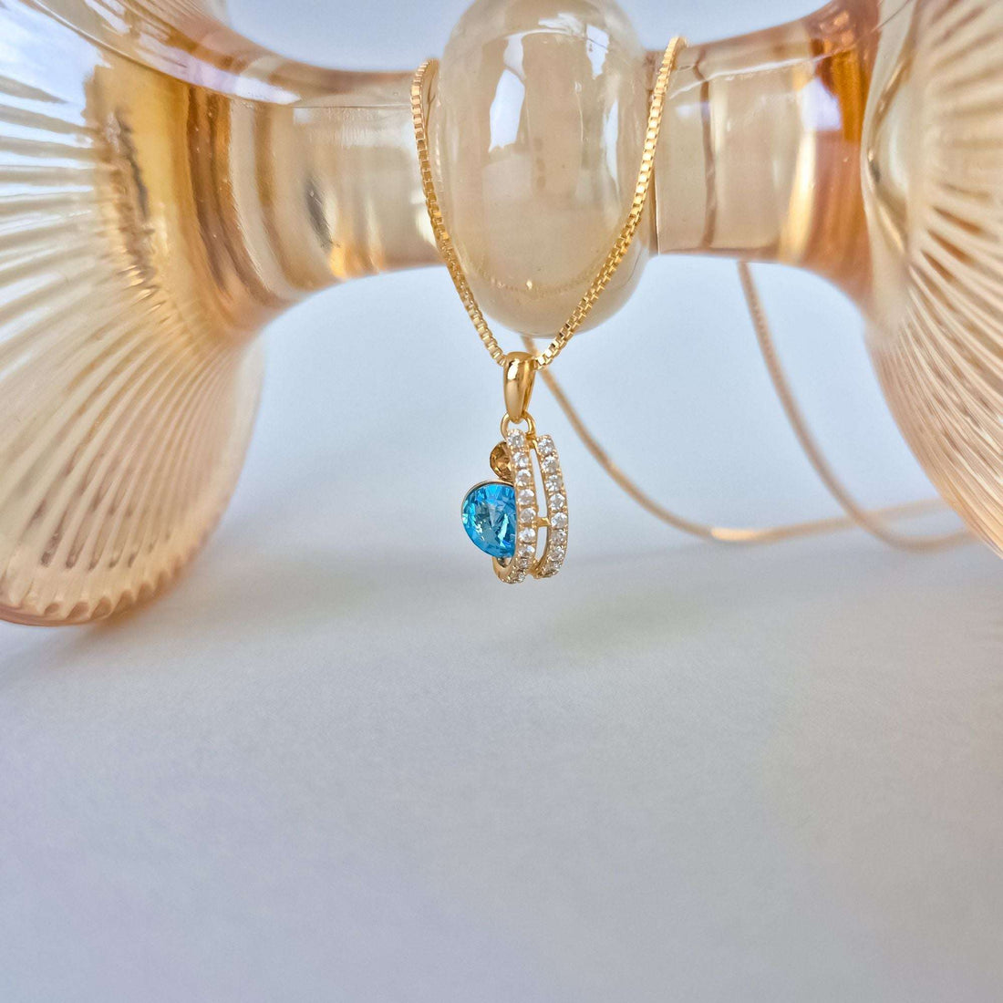 Blue crystal pendant in a gold-tone cubic zirconia frame beside a glass vase