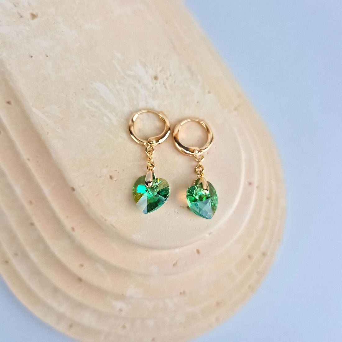 Green heart earrings with round gold-plated hooks on a sand-coloured stand, light blue background