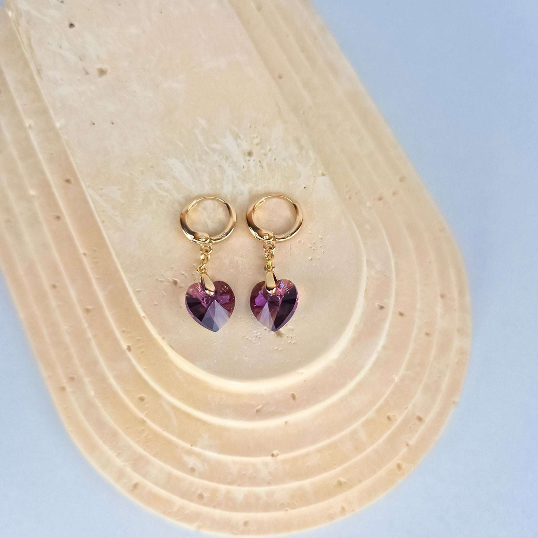 Violet heart crystals hanging from gold-plated hoops on a pale stone stand