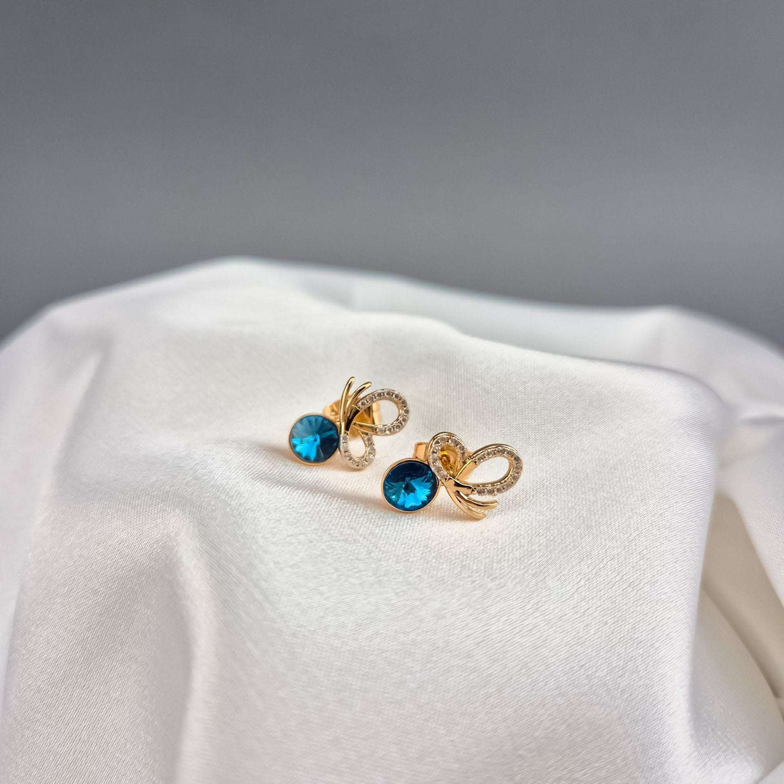 Gold-plated earrings with a blue crystal and bow motif on white satin