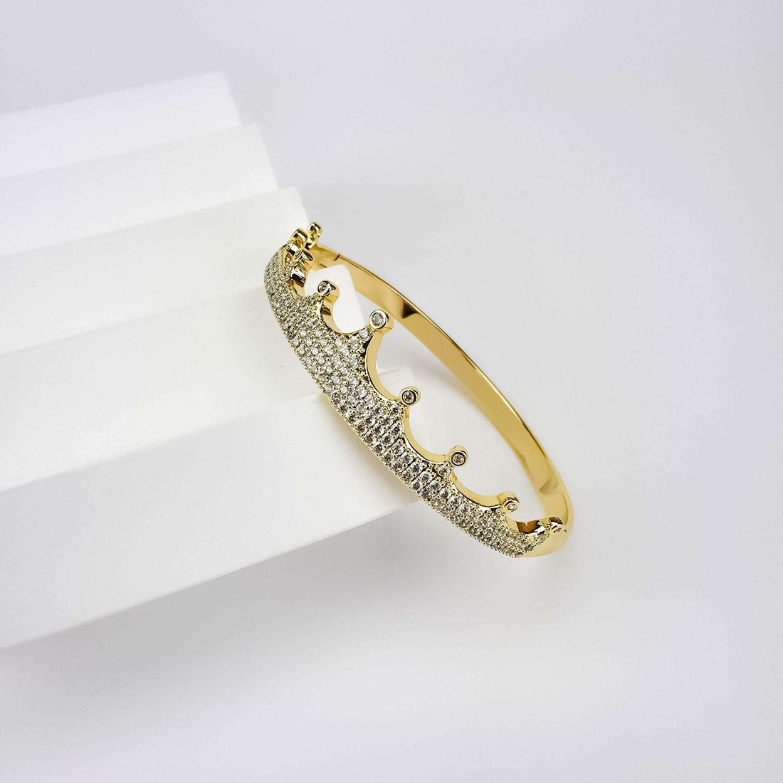 Gold-plated crown bangle at an angle on a white stand, clasp and stones visible