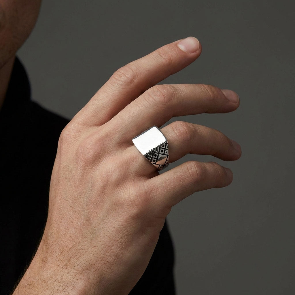 Man's hand wearing a square steel ring with patterned sides, grey background
