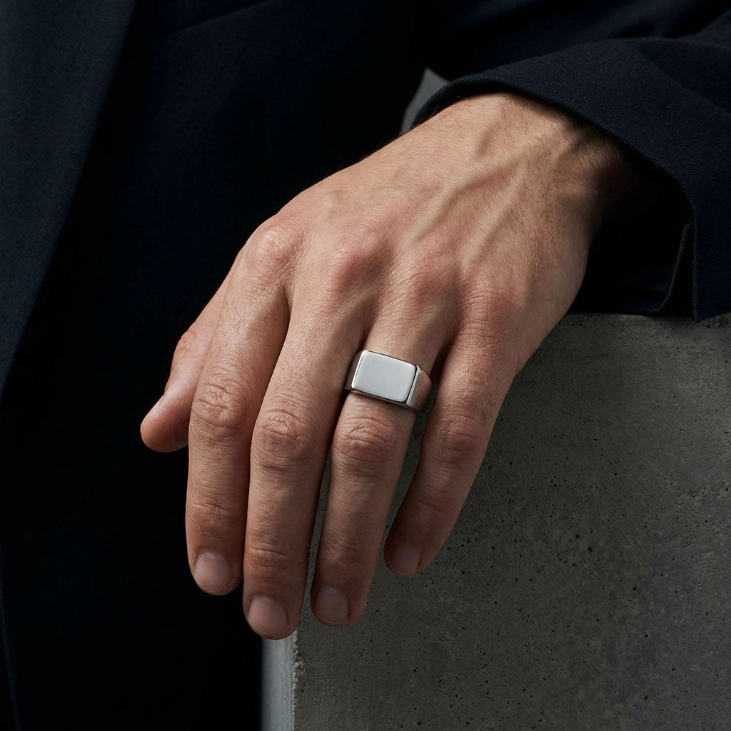 Chunky steel signet ring on a man's finger, hand resting on concrete