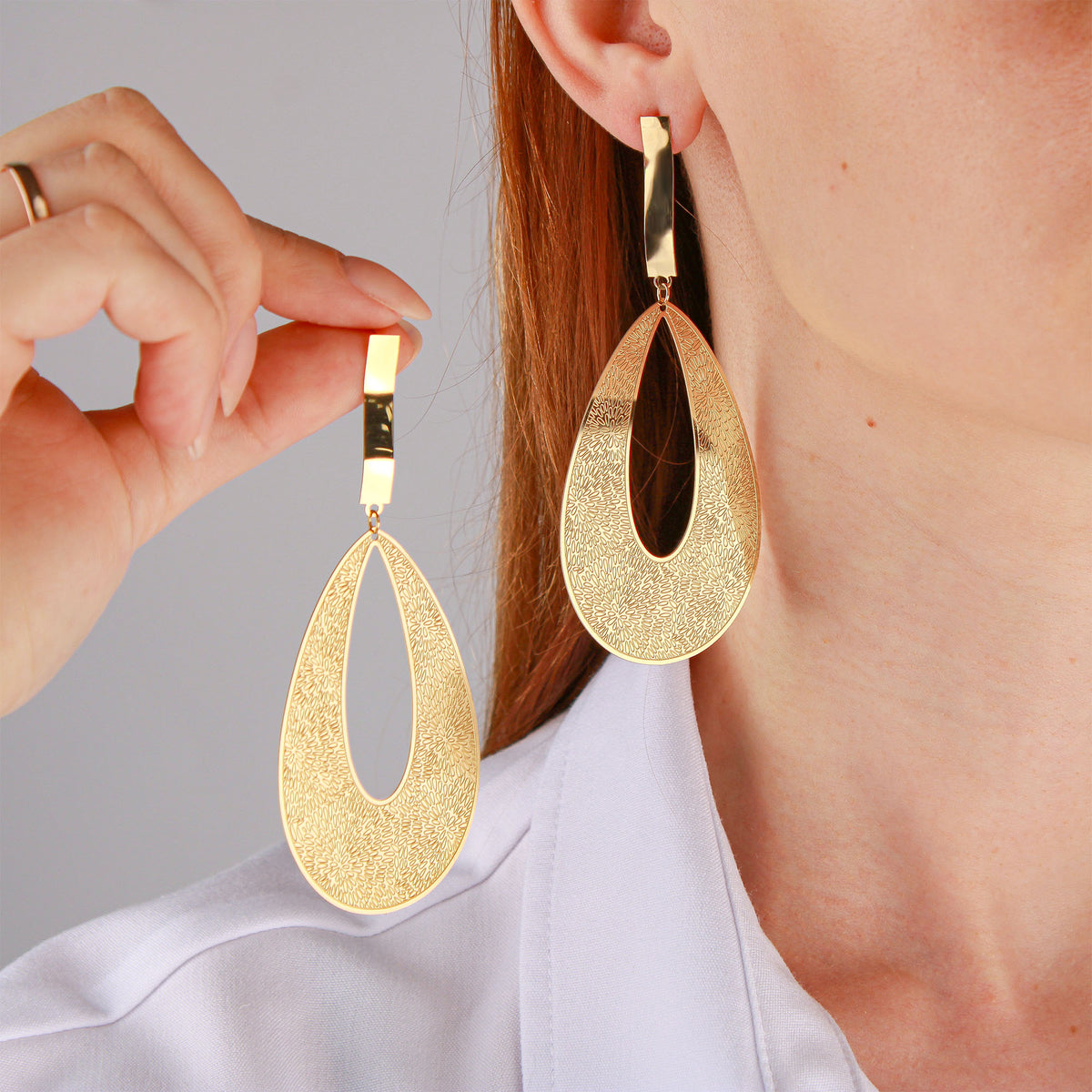 Model in a white shirt wearing and holding large gold-plated teardrop earrings